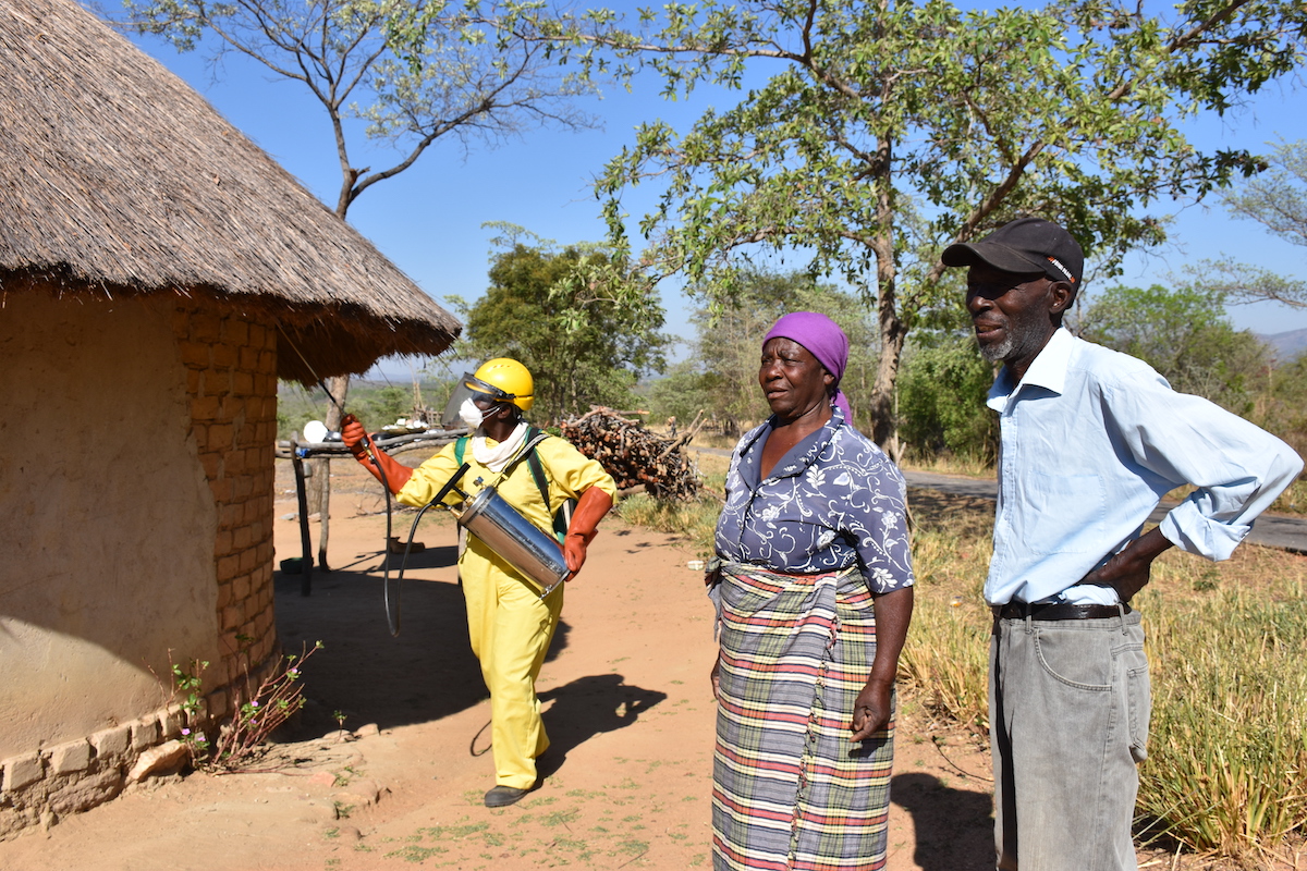 Spray campaign in Zimbabwe showing a rural home with a thatched roof on the left and a spray operator in yellow PPE and a woman with a purple head scarf and a man with a baseball cap on the right side and trees in the background and blue sky behind.