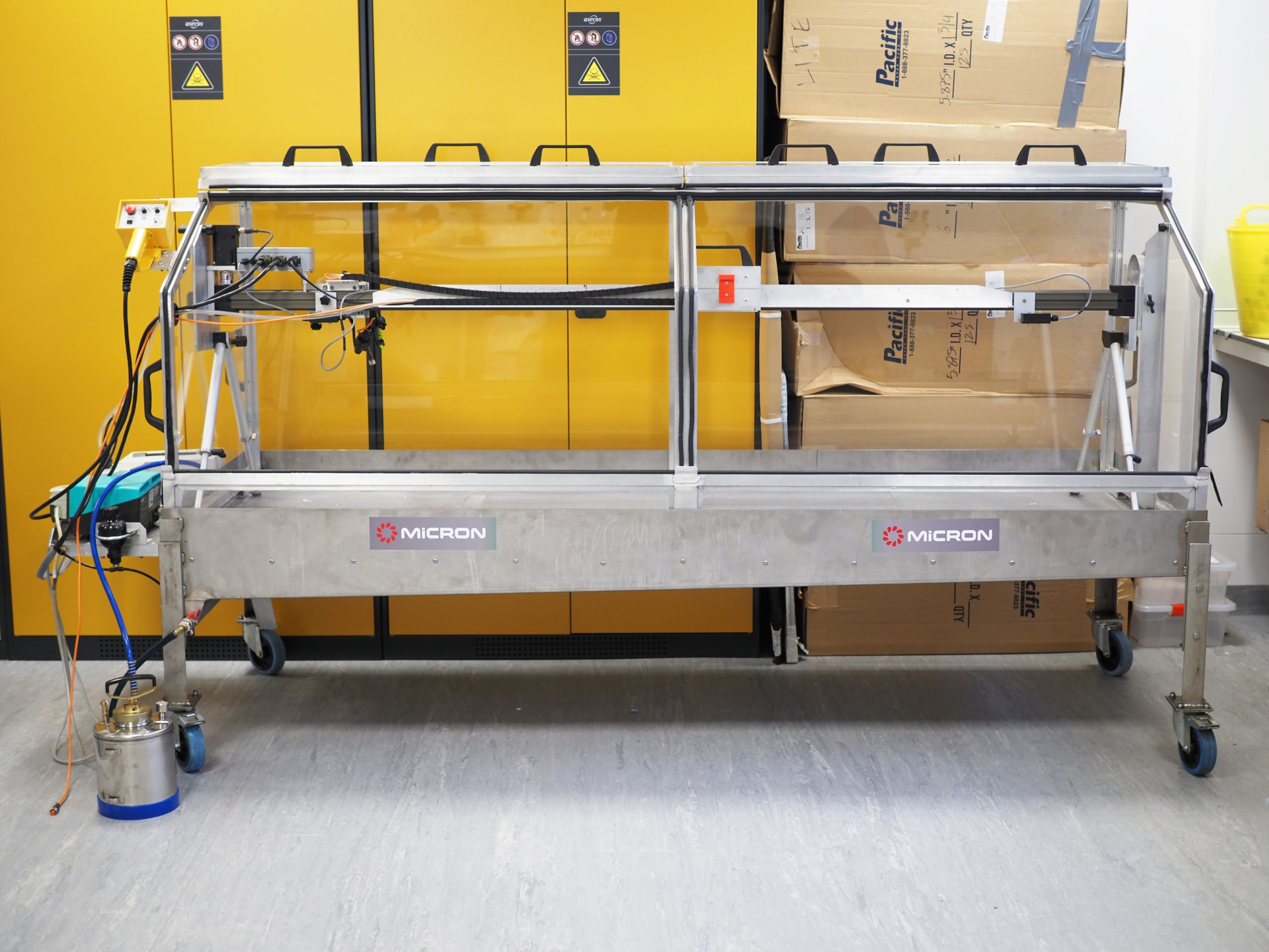 Image of the Track Sprayer. a horizontal, rectangular metal structure with glass doors on all sides with a canister at the base on the left side. Bright yellow cabinets are behind the Track Sprayer.