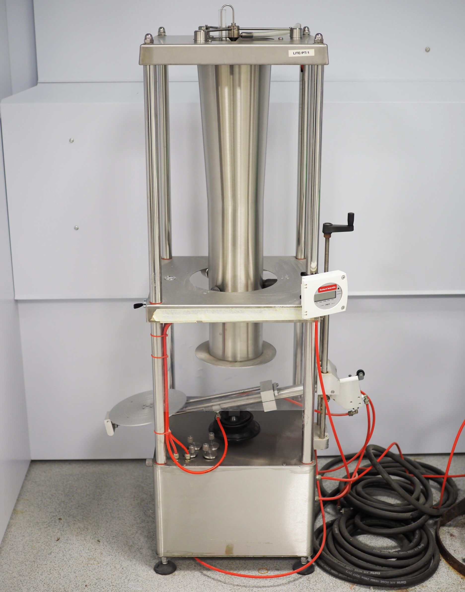 Image of the Potter Tower, a silver, metal rectangular structure with a solid metal base and four metal posts, one on each corner. In the centre is a cylindrical metal funnel that is larger at the top. A metal platform intersects the metal cylinder at the bottom and a revolving handle projects on the right of his platform. Beneath the cylinder is a lever with a crcular platform projecting from the left of the structure. On the floor to the right are coils of black wire and a few red wires are leading into the structure.